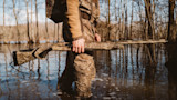Hunter wading through flooded woods, holding camouflaged shotgun horizontally