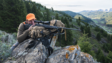 Hunter prone on rocky ridge aiming scoped rifle, orange cap reading FEDERAL, valley below