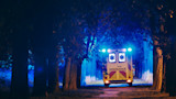 Ambulance with flashing blue lights on a tree-lined path, rear doors read "AMBULANCE"