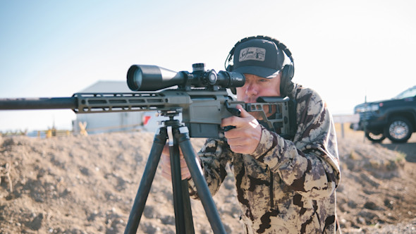 Man aiming scoped rifle mounted on tripod outdoors at a shooting range