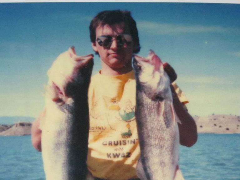 Man holding two large fish, wearing sunglasses and yellow shirt reading "CRUISIN'", on a lake