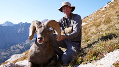 Janis Putelis kneeling beside a bighorn sheep with large curled horns on an alpine slope