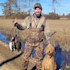 Hunter in camo waders holding a stringer of four ducks and a wet curly dog in flooded field