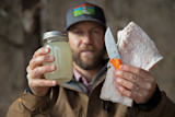 Clay Newcomb holding mason jar of rendered fat and orange-handled knife, hat patch 'Bear Grease'