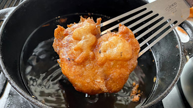 Battered fish fritter on stainless spatula over cast-iron skillet; spatula reads 'Winco Stainless'