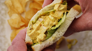Hands holding sandwich filled with curried turkey salad and lettuce on white bread; potato chips behind
