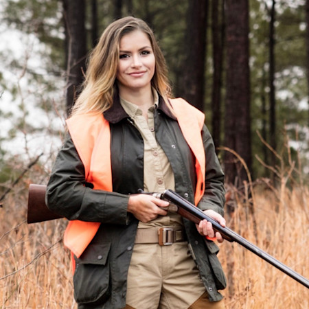Woman wearing orange safety vest holding a double-barrel shotgun in a pine forest