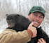 Man holding black bear cub on his shoulder, wearing green cap in snowy woods