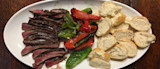 Sliced medium-rare steak, charred red and green peppers, and sliced baguette on white platter