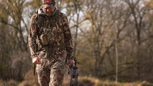 Hunter in full camouflage carrying a compound bow and chest pack, walking through bare woods