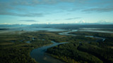Aerial view of winding river through forest with low fog, distant snow-capped mountains and small airplane