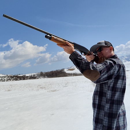 Man aiming shotgun at sky in snowy field, wearing plaid shirt, sunglasses, and trucker cap