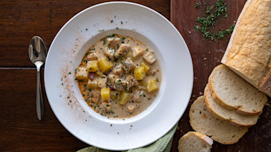 Rockfish chowder with potatoes and herbs in white bowl, sliced baguette and spoon