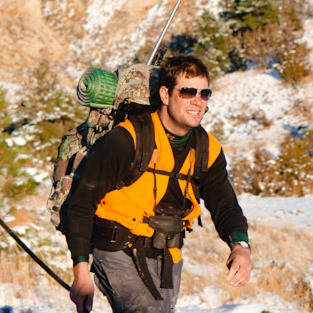 Hunter in orange safety vest and sunglasses carrying backpack, rifle and binoculars in snowy field