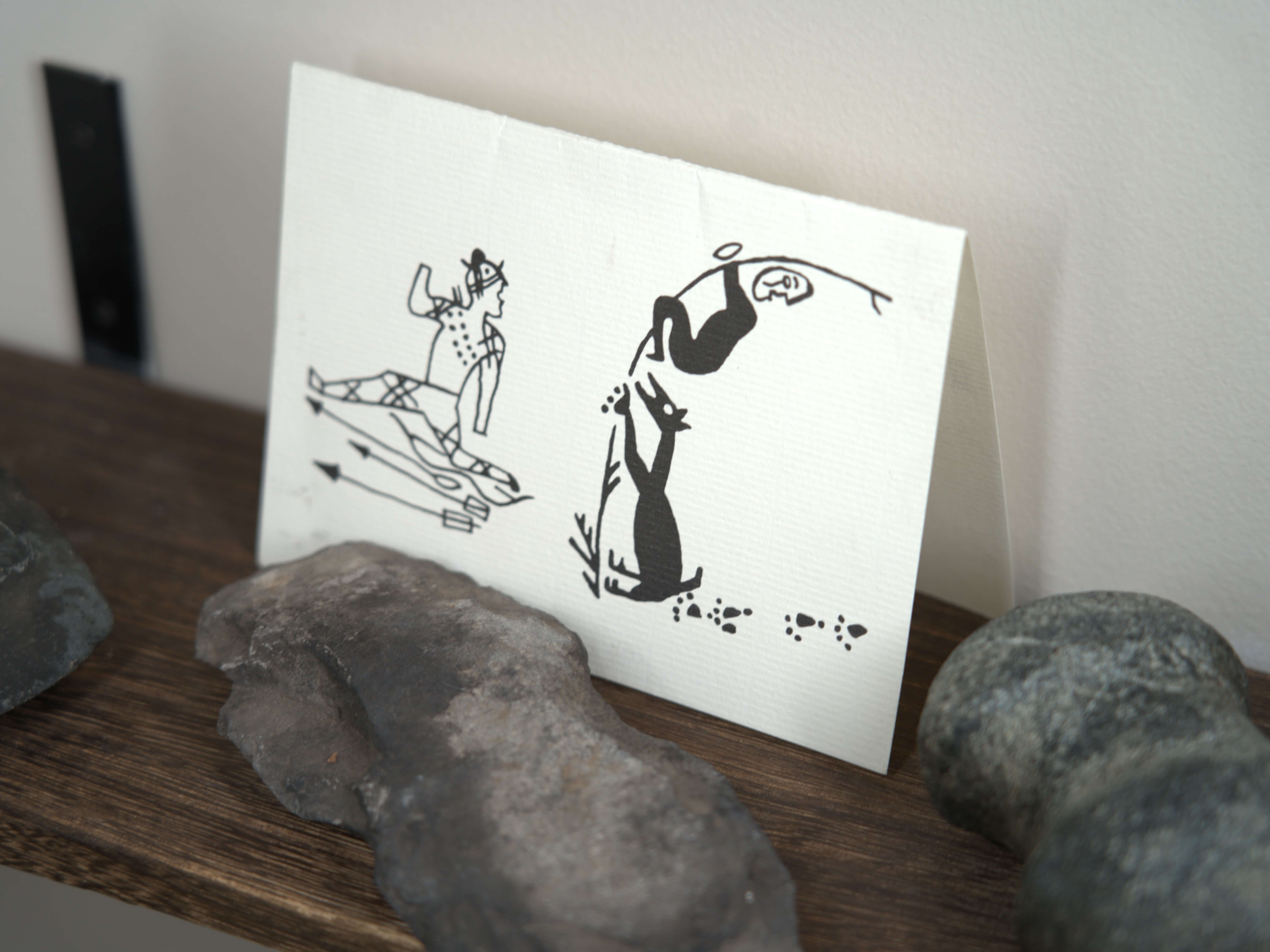 Folded white card with black drawings: person with arrows and fox reaching for fish on a wooden shelf