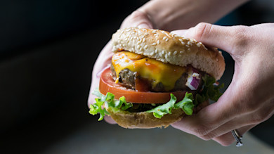 Handheld cheeseburger with sesame bun, lab-grown patty, melted cheddar, tomato, lettuce, red onion