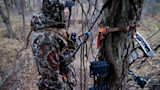 Hunter in camouflage jacket and hood at tree with compound bow, ropes, and binoculars