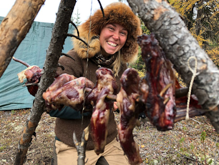 Woman in fur hat smiling while hanging meat on a wooden drying rack outdoors
