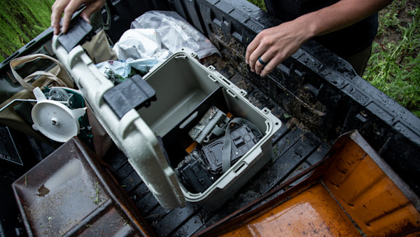 Open waterproof case with trail cameras and batteries on wet truck bed, hands reaching in
