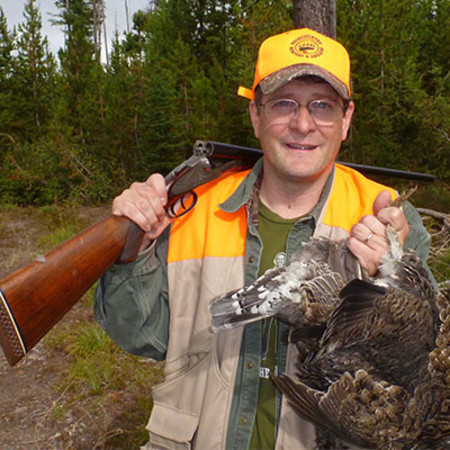 Smiling hunter in orange cap and vest carrying a shotgun and holding game birds
