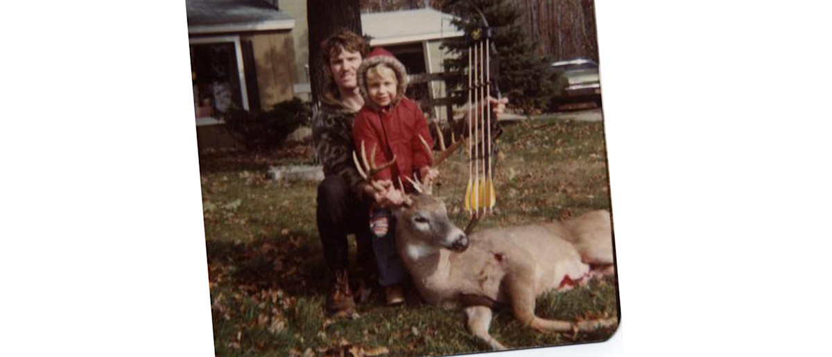 Man kneeling behind child in red coat holding buck antlers; bow and arrows beside deer on lawn