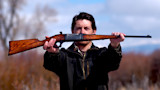Man holding restored wooden-stock rifle horizontally at arm's length in a field