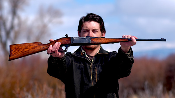 Man holding restored wooden-stock rifle horizontally at arm's length in a field