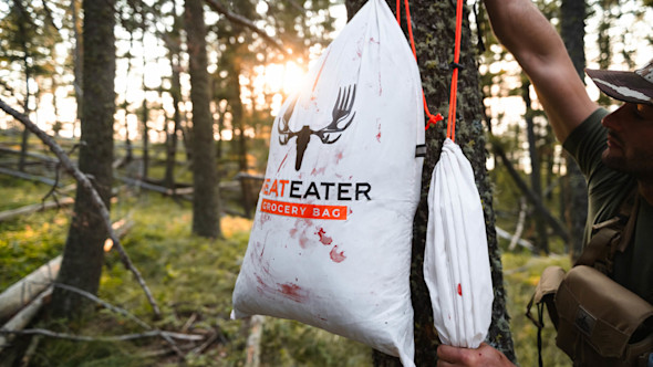 MeatEater grocery bag stained with blood hanging from a tree; man holding orange rope