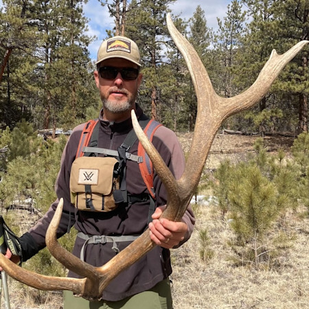 Man wearing sunglasses and cap holding large elk antler in a pine forest