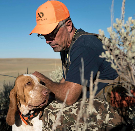 Hunter kneeling, petting brown-and-white hunting dog, wearing orange hat that says 'First Lite'