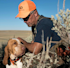 Hunter kneeling, petting brown-and-white hunting dog, wearing orange hat that says 'First Lite'