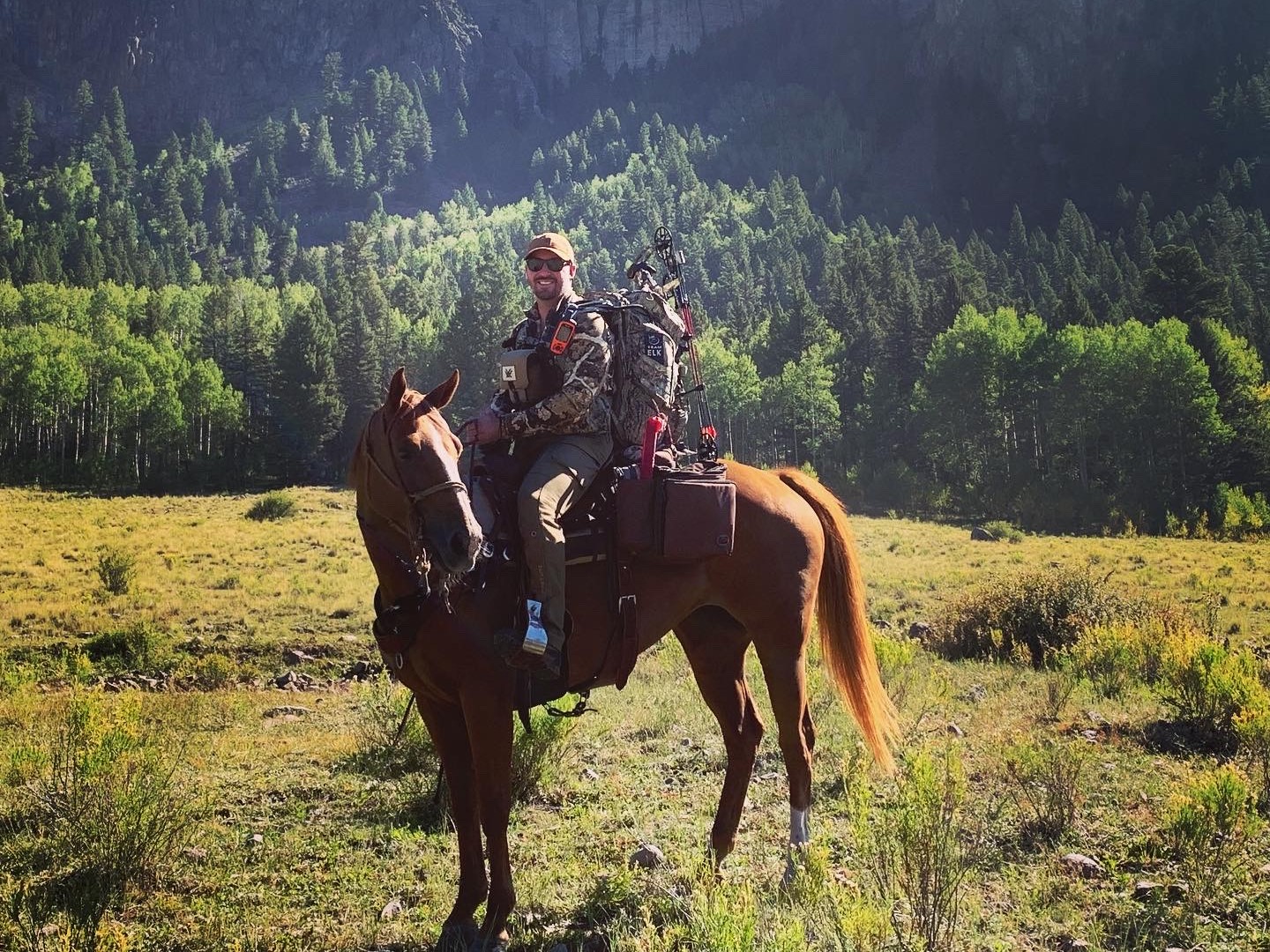 Hunter on horseback with packed gear and compound bow in meadow below forested cliffs