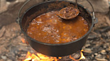 Dutch oven of wild-game chili simmering over campfire, ladle scooping meat and beans