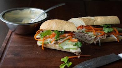 Two banh mi sandwiches with pickled carrots, daikon, cucumber, cilantro and mayo bowl on wooden board