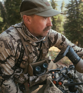 Hunter in camo baselayer with binoculars and First Lite strap, using spotting scope in forest
