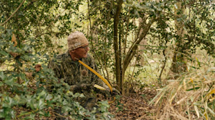 Hunter kneeling in brush holding yellow recurve bow and arrow, wearing camo and tan beanie labeled GHG