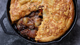 Cast-iron skillet pie with golden puff pastry, slice revealing braised meat and caramelized onions