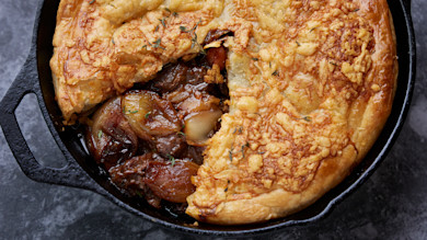 Cast-iron skillet pie with golden puff pastry, slice revealing braised meat and caramelized onions