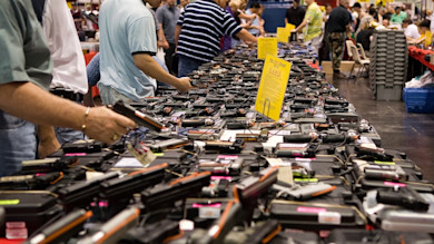 Long table of handguns at a gun show, people browsing; yellow sign reading 'S.A.W.S.'
