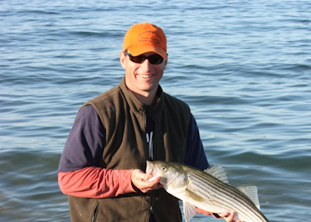 Man standing in water holding a striped bass, wearing an orange cap and sunglasses