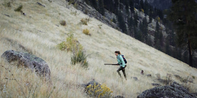 Woman hunter climbing steep grassy hillside with shotgun and backpack, hunting dog behind