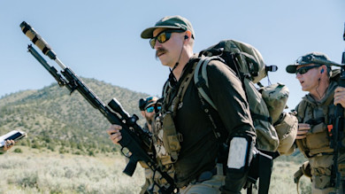 Hunter carrying scoped rifle and large backpack walking in sagebrush with two others