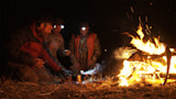 Three hunters kneel by a campfire at night, cooking from a steaming pot with headlamps