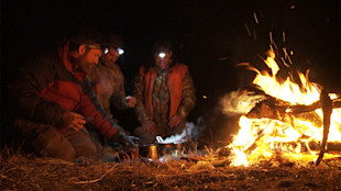 Three hunters kneel by a campfire at night, cooking from a steaming pot with headlamps