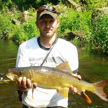 Man holding large golden fish in shallow stream, wearing cap with sunglasses on brim