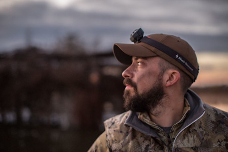Bearded man in camouflage jacket wearing cap and headlamp, side profile at dusk