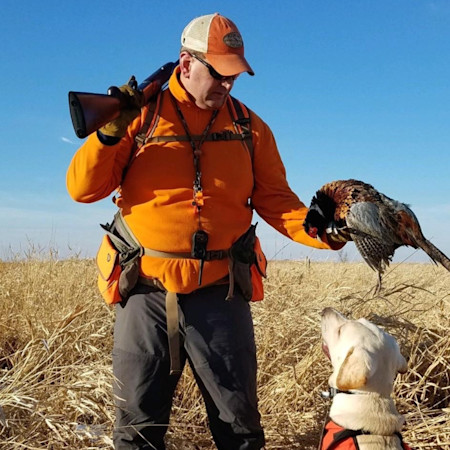 Hunter in orange jacket with shotgun holds a pheasant; dog looks up in grassy field