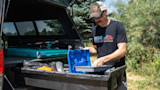 Man at open truck bed organizing blue tackle box, wearing 'RINELLA 2020' shirt and STIHL cap