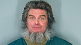 Man with long gray beard and blue eyes against gray wall, mug shot