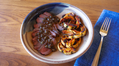 Sliced roast topped with capers and sauce, charred delicata squash spirals in ceramic bowl
